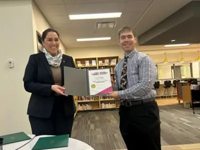 Two people standing in the school library holding and award between them. On the left is Member of Parlaiment Sima Acan wearing a black suit, brown shirt and a beige scarf, on the right is Vittorio Iafrate, winner of the Prime Ministers Teaching Excellence Award. He is wearing a blue striped shirt and a black and yellow tie and black pants. There are two green folders on the round table in front of them.