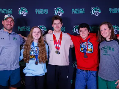 Male coach on the left side wearing greay shirt and blue shorts. Girl in blue shirt with medal around neck, boy in the middle has three medals with red and blu ribbons wearing beig shirt, boy in the middle left has medal on blue ribbon wearing red shirt. Female coach on far right hand side. wearing great and black shirt that says "Blakelock athletics."