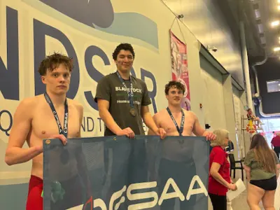3 student members of the swim team are standing in front of a large blue flag that says OFSAA. The boy on the left is wearing red shorts and no shirt he has a blue ribbon around his neck. The boy in the middle is wearing a grey "Blakelock" tshirt and a medal around his neck that has a blue ribbon. The third boy is on the right hand side of the photo is wearing no shirt and also has a blue ribbon around his neck. They won medals for swimming.