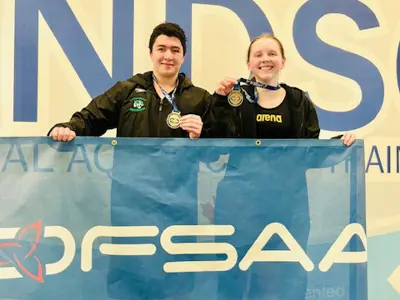 Two students one male on the left and one female on the right standing in front of a large blue flag that says OFSAA. They are both smiling and holding gold medals for swimming.