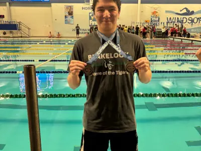 Male blakelock student is standing in front of the pool holding tso medals that he has around his neck both on blue ribbons. He is smiling. There are some people way behind him on the other side of the pool.