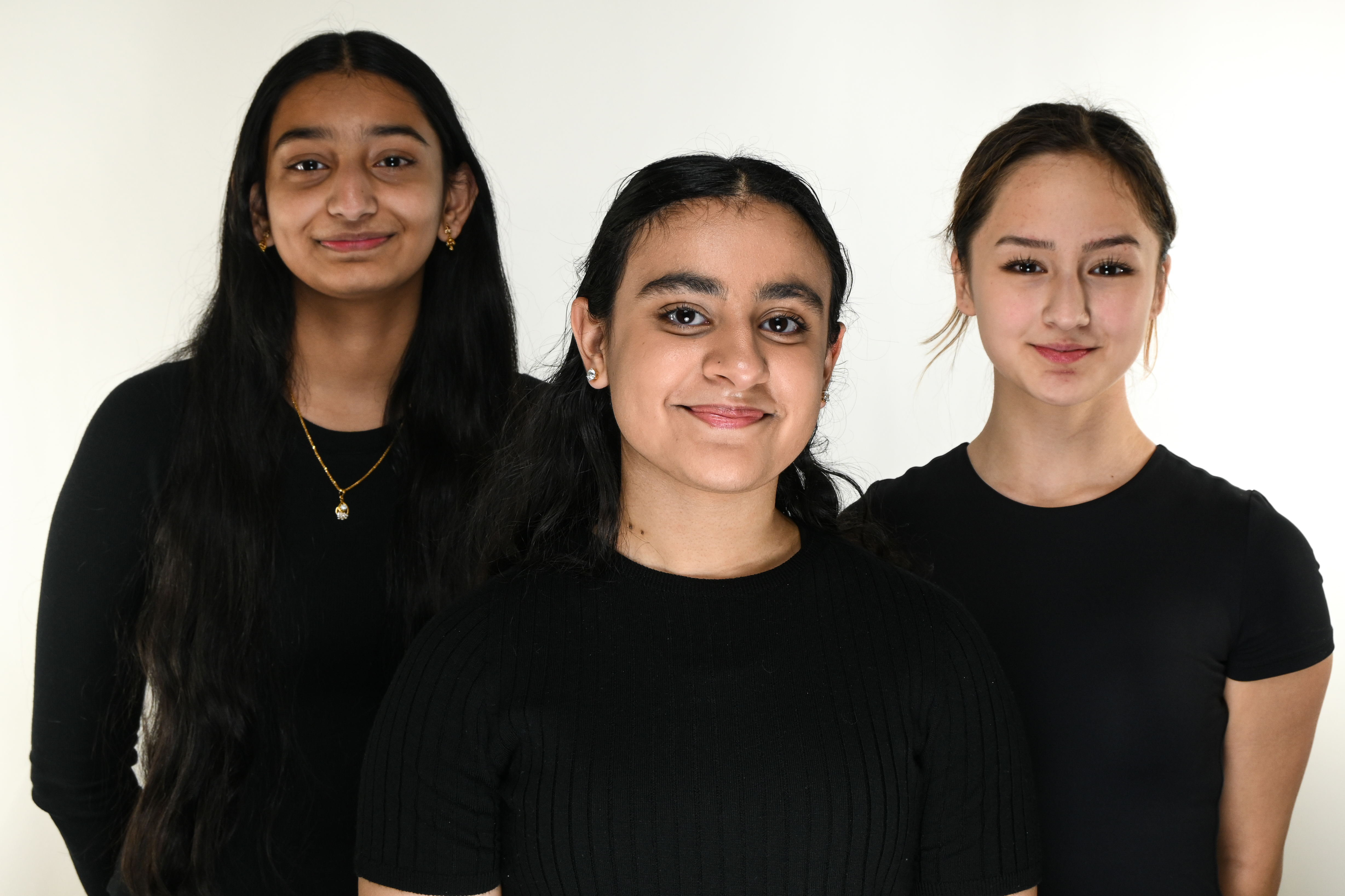 Three high school students, Emmy Nah, Amrutha Kosuri and Veera Kalsi, stand together smiling, wearing black shirts against a white background.