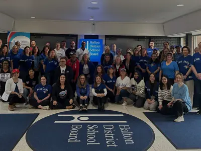 Group of Halton District School Board staff in the lobby wearing Toronto Blue Jays shirts and jerseys, smiling and posing around the HDSB floor logo and sign.