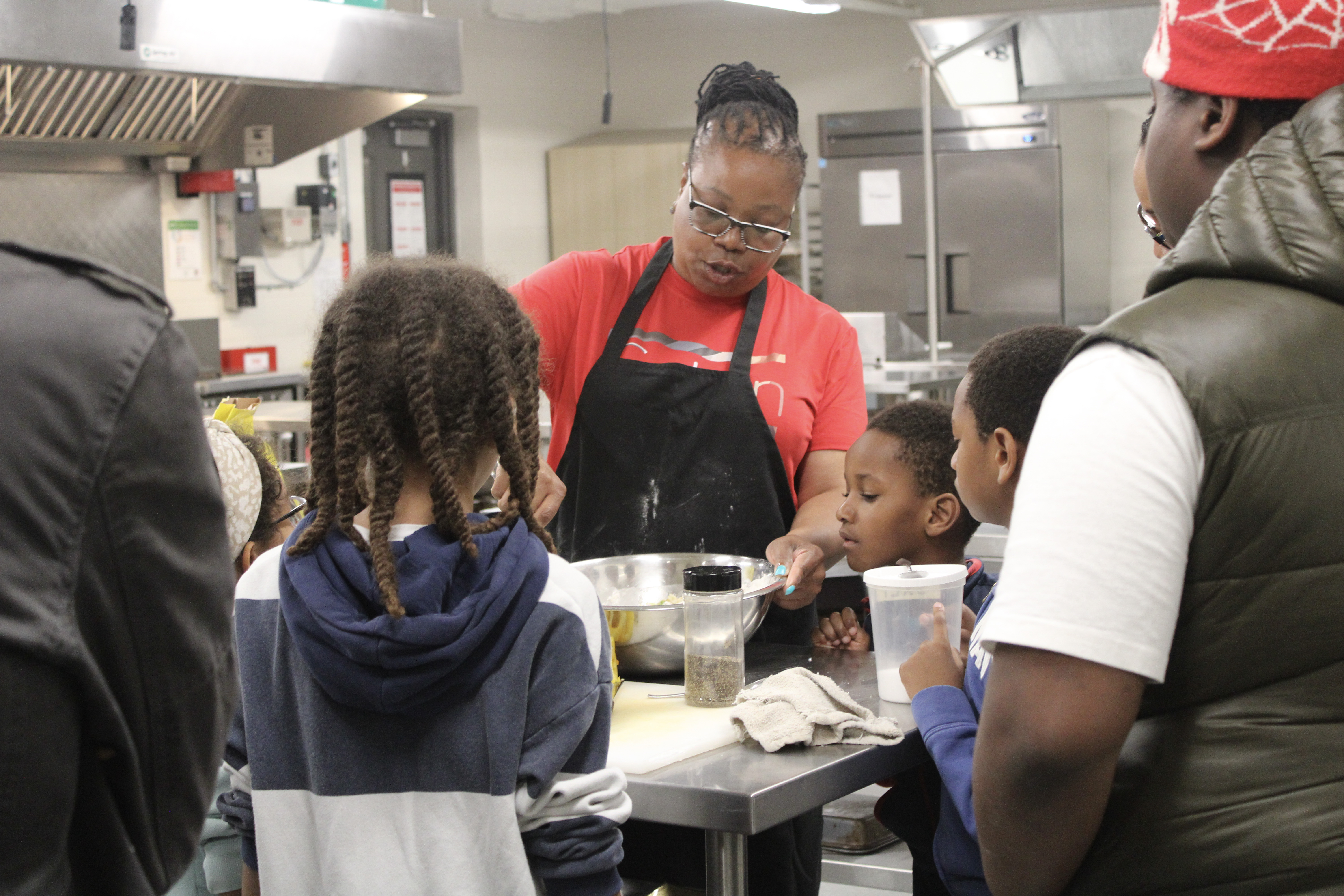 A teacher in a red shirt and black apron leads a hands-on cooking lesson for a group of students gathered around a kitchen workstation in a school setting.
