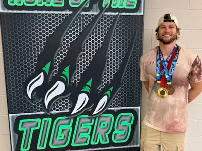 Paralympic Gold medalist Austin Smeenk is standing on the right hand side of the photo next to the large green and black sign that says "Home of the Tigers" with the big claw mark on it. He is smiling and wearing beige pants and a beige shirt as well as his three medals from the ParaOlympics. On medal is on a red ribbon and two are on blue ribbons around his neck. He is also wearing a beige ball cap on backwards.