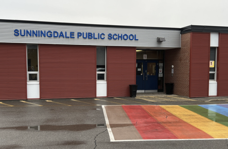 The front entrance of Sunningdale Public School, featuring red corrugated siding, a blue door labeled "1", and a large rainbow-painted crosswalk on the pavement in the foreground.