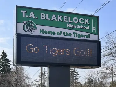 A large digital sign outside. The top half is printed and says "T.A.Blakelock High School Home of the Tigers!" and the bottom half is a changing digital sign that in the currently in the photo says "Go Tigers Go!!!" There are trees in the background at the bottom of th photo.