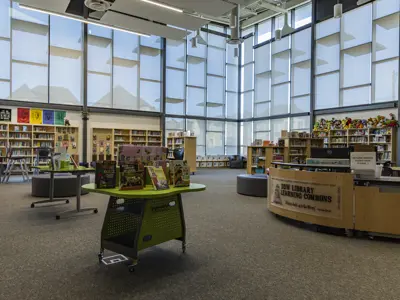 A bright, modern school library with tall floor-to-ceiling windows letting in natural light. Bookshelves line the walls, and colorful books are displayed on round tables in the center. Soft seating stools and chairs are scattered throughout the open space for reading. On the right, a circulation desk labeled “DDW Library Learning Commons” sits near a return bin for library books. Stuffed toys decorate the top of a bookshelf, adding a playful touch to the welcoming learning environment