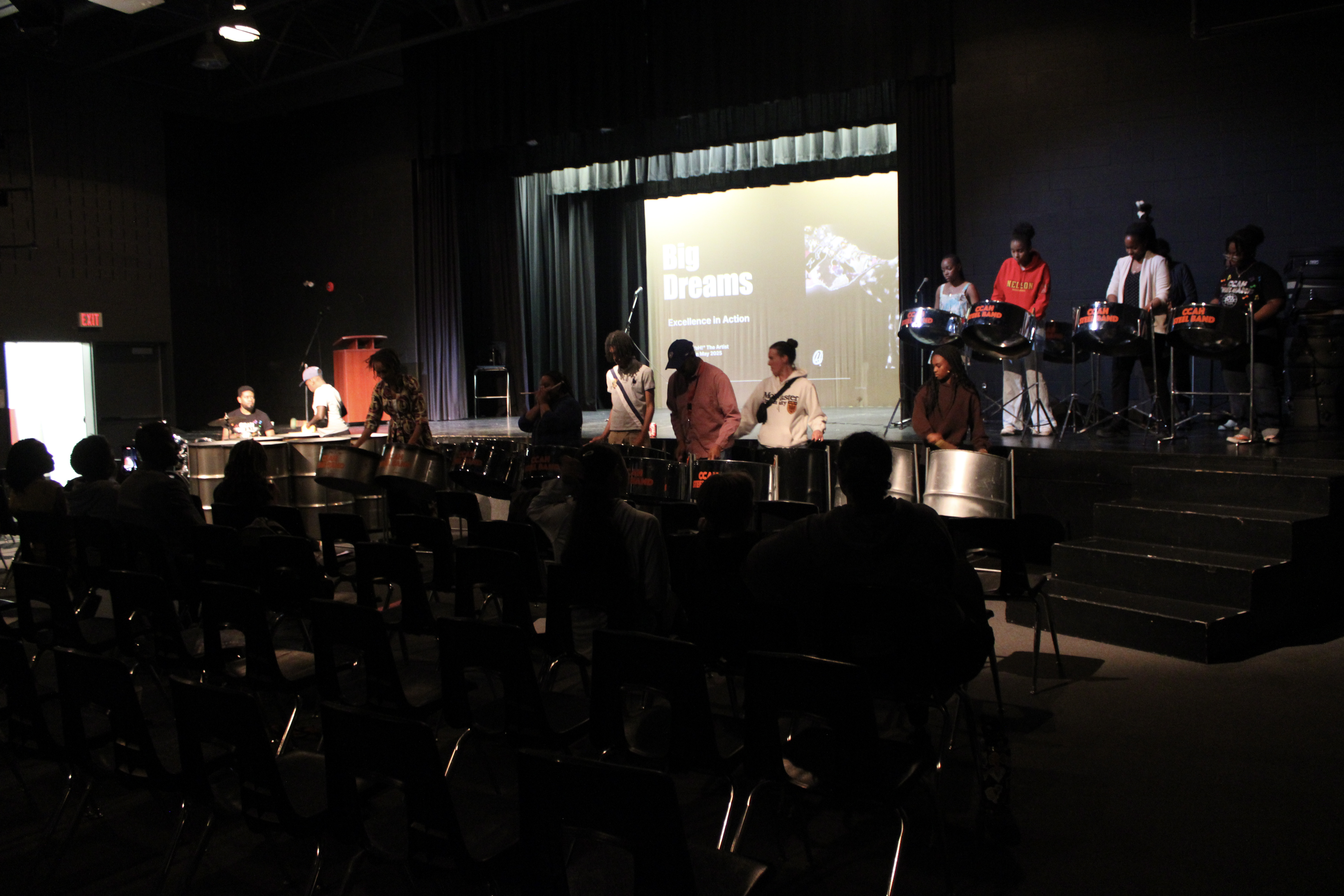 Students performing on steel drums on a dimly lit stage with a screen in the background displaying 'Big Dreams'.